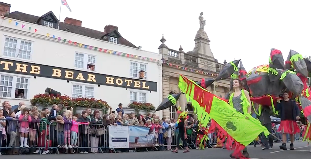 a large spider carnival float makes its way through Devizes Market Place with an audience watching